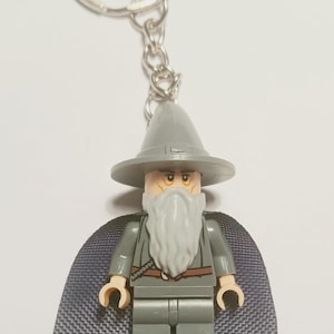 Op de afbeelding: Een sleutelhanger met een plastic minifiguur van een tovenaar met een grijze puntmuts, een lange grijze baard en een grijs gewaad. Het figuurtje draagt een donkergrijze stoffen cape en zit met een metalen kettinkje aan een zilverkleurige ring.