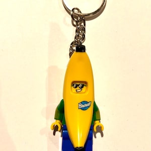 Puede incluir: Un llavero con una minifigura de plástico que lleva un disfraz de plátano amarillo. La figura tiene una cara amarilla con gafas de sol negras y una sonrisa blanca. Un logotipo circular azul con el texto BANANA! aparece en el disfraz. La figura lleva mangas verdes y pantalones azules, y está unida a una anilla de metal plateado.