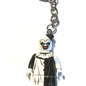Könnte beinhalten: Ein Schlüsselanhänger mit einer kleinen Kunststofffigur, die als Clown mit weißem Gesicht, schwarzen Augenmarkierungen und einem breiten Grinsen gestaltet ist. Die Figur trägt ein schwarz-weißes Outfit mit Rüschenkragen und einem kleinen schwarzen Hut.