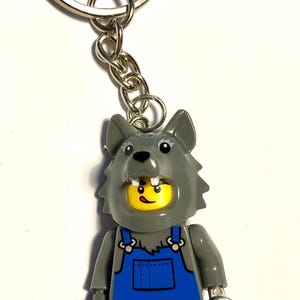 Może przedstawiać: Brelok z plastikową minifigurką w szarej masce wilczej głowy z widocznymi białymi zębami. Minifigurka ma żółtą twarz z uśmieszkiem i jest ubrana w niebieskie ogrodniczki na szarym korpusie. Dołączone jest srebrne metalowe kółko z łańcuszkiem.