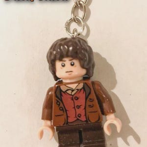 Op de afbeelding: Een sleutelhanger met een klein plastic figuurtje met bruin haar, een bruin jasje, een rood vest en een donkerbruine broek. Het figuurtje heeft een neutrale gezichtsuitdrukking met zwarte ogen en wenkbrauwen. De tekst Brick Chica is linksboven zichtbaar.