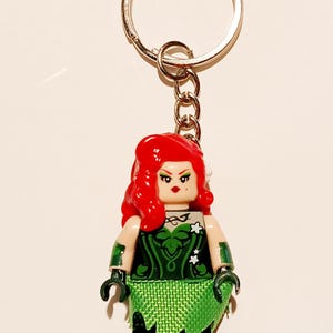 Könnte beinhalten: Ein Schlüsselanhänger mit einer Miniaturfigur mit leuchtend rotem Haar, einem grünen Oberteil mit Blattmustern und einem strukturierten grünen Rock. Die Figur hat dunkelgrüne Armstulpen und schwarze Beine und ist an einem silberfarbenen Ring befestigt.
