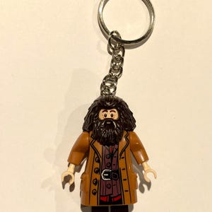 Può includere: Un portachiavi con una minifigure in plastica di un personaggio con lunghi capelli castano scuro e una folta barba. La figura indossa un lungo cappotto marrone sopra una camicia rosso scuro con cintura nera e fibbia, e pantaloni marrone scuro.