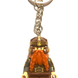 May include: A keyring featuring a plastic minifigure of a dwarf with a long orange beard, wearing a brown helmet with gold detailing, a brown tunic with a gold belt buckle, and grey sleeves. The figure is attached to a silver metal ring.