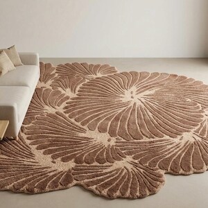 May include: A large, irregularly shaped area rug featuring a textured, repeating fan-like pattern in shades of brown and cream. The rug has a thick, plush pile and covers a significant portion of the floor, extending underneath a light-colored sofa.