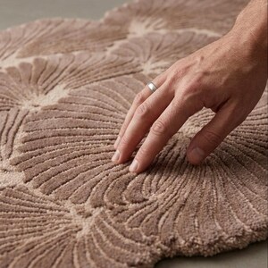 May include: A tufted rug in a muted taupe color featuring a repeating fan-shaped or shell-like pattern. The surface has a textured, raised pile construction with deep grooves between the curved segments, creating a tactile and dimensional appearance.