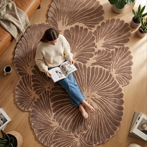 May include: A large, irregularly shaped area rug in a warm taupe color, featuring a repeating fan-like or shell-inspired embossed pattern across its textured surface. The rug covers a significant portion of the floor, showcasing a soft, plush pile.