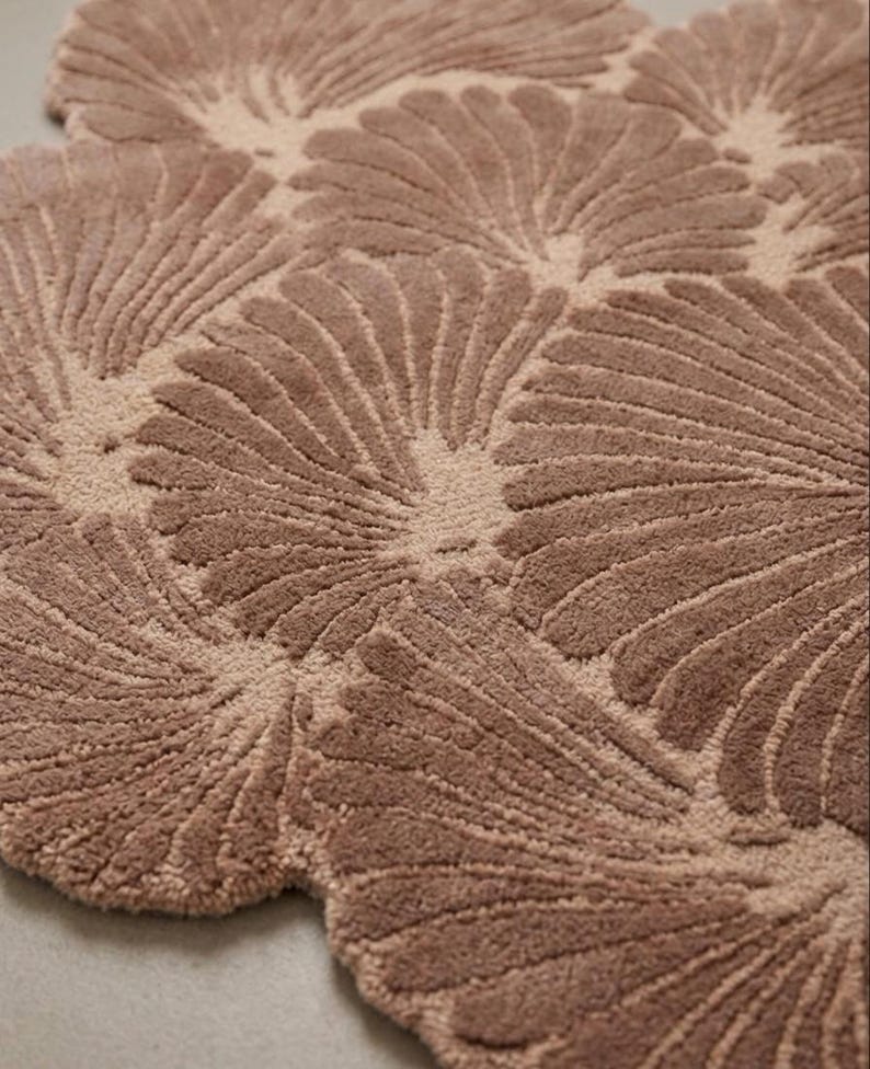 May include: A tufted area rug in a dusty rose color featuring a repeating pattern of fan-shaped or shell-like motifs. The design consists of raised, textured lines radiating from a central point within each shape, creating a soft, plush surface.