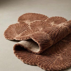 May include: A brown bath mat with a textured, repeating fan-shaped pattern. The rug features a soft, tufted pile surface and a folded edge that reveals a light-colored, woven fabric backing. The overall shape is organic, composed of multiple rounded sections.