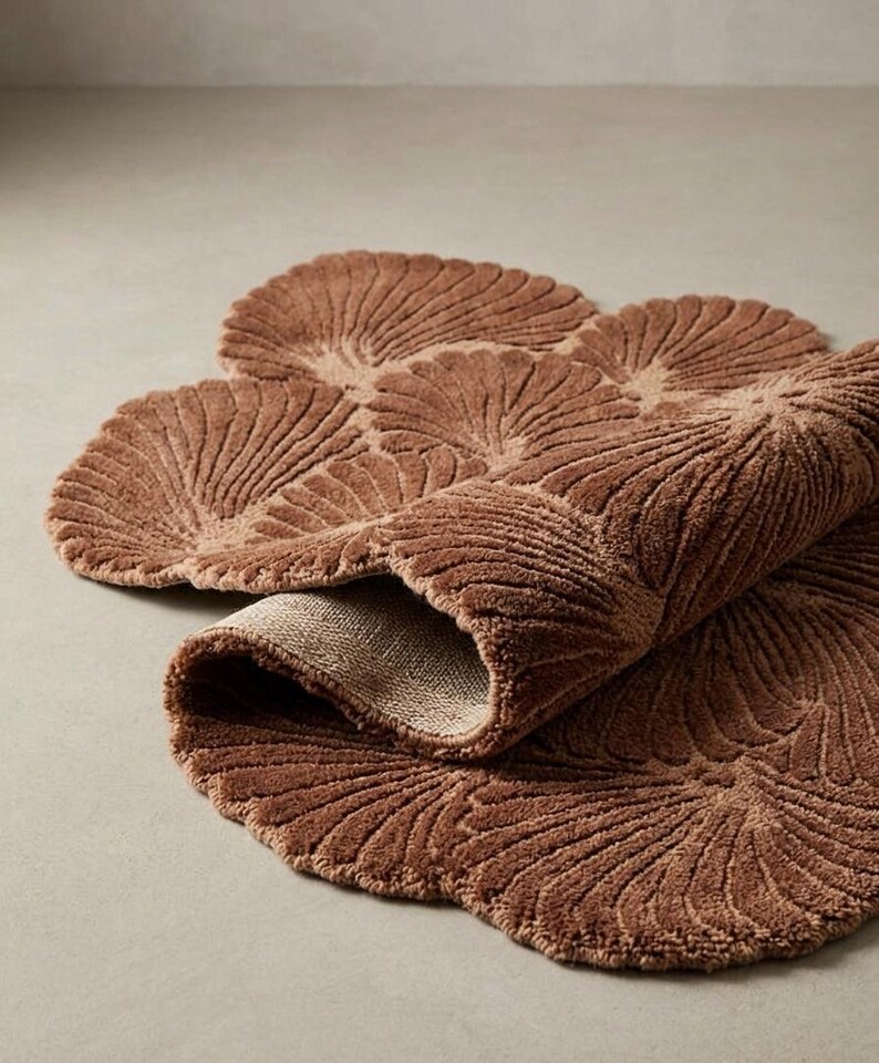 May include: A brown bath mat with a textured, repeating fan-shaped pattern. The rug features a soft, tufted pile surface and a folded edge that reveals a light-colored, woven fabric backing. The overall shape is organic, composed of multiple rounded sections.