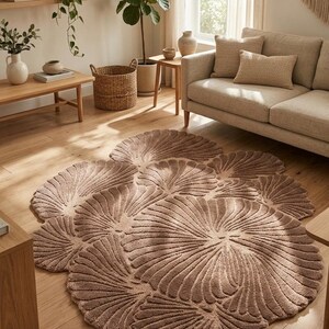 May include: A large, circular area rug in a muted taupe color, featuring a textured, raised pattern of overlapping fan-shaped or shell-like segments that radiate outward from the center, creating a distinct, organic, and layered floral silhouette.