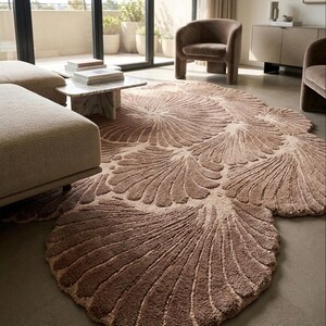 May include: A large, irregularly shaped area rug in a muted taupe color, featuring a repeating pattern of raised, fan-like shell motifs with deep grooves. The rug has a thick, textured pile and covers a significant portion of the floor space.