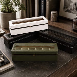 May include: A collection of four rectangular watch display cases with clear glass lids and internal compartments. The boxes are finished in white, black, and olive green, each featuring a central metal latch closure and visible stitching along the edges.