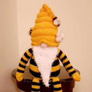 Könnte beinhalten: Ein gestrickter Bienengnom mit einem gelb-schwarz gestreiften Körper, einem weißen Bart und einem gelben Honigwabenhut mit weißen Blüten. Der Gnom sitzt auf einem Holztisch. "Vorderansicht" steht in der rechten unteren Ecke des Bildes.