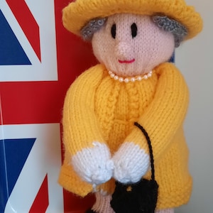 May include: A knitted doll wearing a yellow dress, hat, and white gloves. The doll has a pink face with black eyes and a pearl necklace. The doll is holding a black purse and is standing in front of a Union Jack tray.