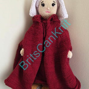 May include: A handmade knitted doll wearing a red cloak and a white bonnet. The doll has a light skin tone, black eyes, and a small pink mouth. The doll's feet are visible, wearing gray and yellow shoes. The text "BritsCanKnit" is visible on the image.
