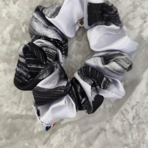 Puede incluir: Un coletero de tela con un patrón abstracto en blanco y negro y un acabado suave similar al satén. El texto "Chouchou 7" está impreso sobre el artículo. Una pequeña etiqueta con rayas azules, blancas y rojas es visible en el borde inferior.