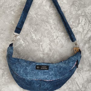 Puede incluir: Un bolso bandolera en forma de media luna hecho de tela azul con aspecto de mezclilla y una correa larga ajustable. El bolso presenta una etiqueta rectangular negra con el texto blanco L'atelier de servane y clips de metal plateado.