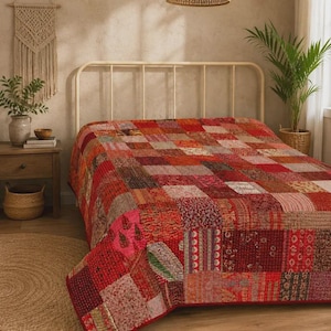Puede incluir: Una colcha de patchwork que presenta una variedad de cuadrados de tela rojos, rosas y naranjas con diversos patrones florales y geométricos. La colcha está extendida sobre una estructura de cama de metal blanco con costuras visibles.