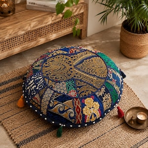 May include: A round floor cushion featuring a patchwork design of blue, green, and gold fabrics with intricate embroidery, metallic thread detailing, and small sequins. The cushion is accented with colourful tassels in red, yellow, and green.