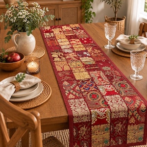 K&ouml;nnte beinhalten: Ein rechteckiger Tischl&auml;ufer mit einem Patchwork-Design aus verschiedenen roten, goldenen und bunten Stoffquadraten mit aufwendigen Stickereien, metallischen Fadenverzierungen und kleinen Spiegelakzenten, flach auf einem Holztisch ausgelegt.