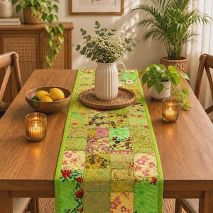 Könnte beinhalten: Ein rechteckiger Tischläufer mit einem Patchwork-Design aus verschiedenen grünen, gelben und floral gemusterten Stoffquadraten. Der Läufer hat einen einfarbigen lindgrünen Rand und durchgehende Steppnähte auf der gesamten Oberfläche.
