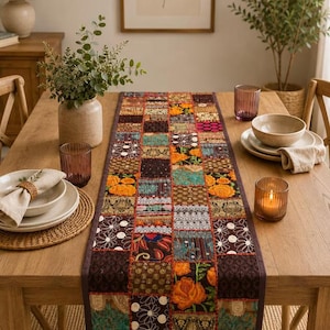 Può includere: Un runner da tavola rettangolare caratterizzato da un design patchwork composto da vari quadrati di tessuto nei toni dell'arancione, marrone, verde acqua e crema. Il runner presenta motivi floreali, forme geometriche e ricami.