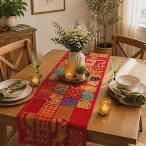 Puede incluir: Un camino de mesa rectangular con un diseño de patchwork de cuadrados de tela rojos, dorados y multicolores con patrones intrincados. El camino está colocado sobre una mesa de madera con platos blancos, servilletas verdes y portavelas de cristal.