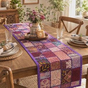 Pode incluir: Um caminho de mesa retangular com um design de patchwork de vários quadrados de tecido em tons de roxo, rosa e dourado. O caminho tem uma borda roxa sólida e exibe bordados intrincados, padrões florais e motivos geométricos.