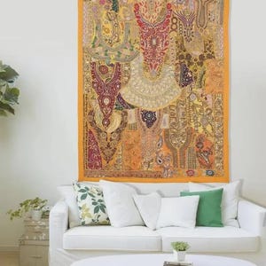 Op de afbeelding: Een rechthoekig wandkleed bestaande uit een patchwork van gele, oranje en rode stoffen panelen. Het textiel is versierd met ingewikkeld borduurwerk, accenten van metaaldraad en kleine ronde spiegeltjes. Het heeft kwastjes aan de bovenhoeken.