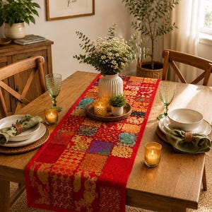 May include: A rectangular table runner featuring a patchwork design of various red, orange, and gold fabric squares with intricate floral and geometric patterns. The runner is centred on a wooden dining table with place settings and a white vase.