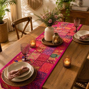 May include: A rectangular table runner featuring a vibrant patchwork design in shades of pink, magenta, and purple with intricate floral and geometric patterns. The runner is laid across a wooden dining table, accented by glassware and candles.