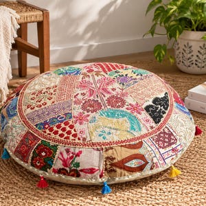 May include: A round floor cushion featuring a patchwork design of various fabrics in red, blue, green, and cream tones. The surface is decorated with intricate floral and geometric embroidery, finished with small colorful tassels along the edge.