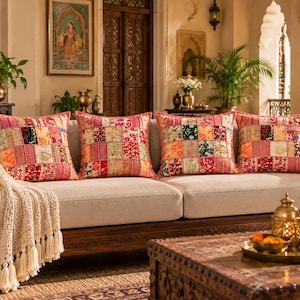 May include: Four square throw pillows featuring a vibrant patchwork design with various floral and geometric patterns in shades of pink, red, orange, and green. The pillows are arranged in a row on a light-colored sofa with a cream-colored throw blanket.
