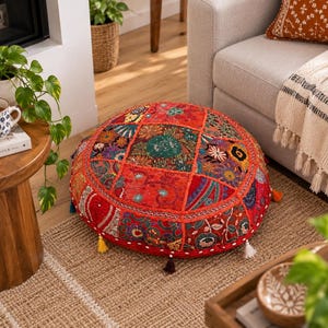 May include: A round floor cushion featuring a patchwork design of vibrant red, orange, green, and blue fabrics. The surface is decorated with intricate embroidery, floral patterns, and small mirrors, finished with colourful tassels along the edge.
