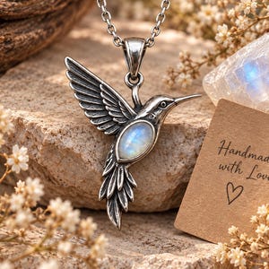 Peut inclure: Un pendentif colibri argenté avec une pierre de lune ovale incrustée sur le corps de l'oiseau, suspendu à une chaîne en argent. Une petite carte beige porte l'inscription Handmade with Love accompagnée d'un symbole de cœur dessiné en dessous.