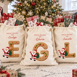 Pode incluir: Três sacos de presente de lona cor de creme com cordão, com o texto vermelho SPECIAL DELIVERY FOR seguido pelos nomes Evelyn, Gloria ou Lincoln. Cada saco exibe uma letra grande de pão de mel E, G ou L com um Papai Noel.