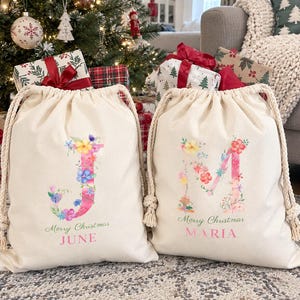Pode incluir: Duas sacolas de presente em lona off-white com cordão, apresentando letras J e M com padrões florais. Cada sacola exibe o texto Merry Christmas em letra verde e um nome personalizado em letras maiúsculas rosas, JUNE e MARIA.