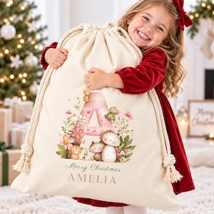 Pode incluir: Um saco de presente de tecido cor de creme com cordão, apresentando uma letra A grande rosa decorada com ilustrações florais e pequenos animais. O texto Merry Christmas e o nome AMELIA estão impressos em rosa na parte frontal.