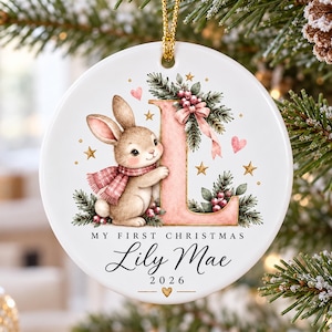 May include: A round white ceramic Christmas ornament featuring a brown bunny wearing a pink scarf next to a large pink letter L decorated with pine branches and berries. The text reads MY FIRST CHRISTMAS Lily Mae 2026 with a small heart.