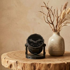 May include: A black, circular, 3D-printed spotlight-style lamp featuring a bat-shaped cutout on its front grille. The lamp is mounted on a circular base with a pivoting mechanism, allowing for adjustable positioning of the light housing.