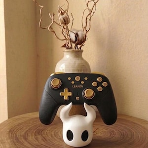 May include: A black wireless video game controller with gold-coloured buttons, joysticks, and d-pad, featuring the text LEA&JDY. The controller rests on a white, horned character-shaped stand with two large black oval eye-like shapes.