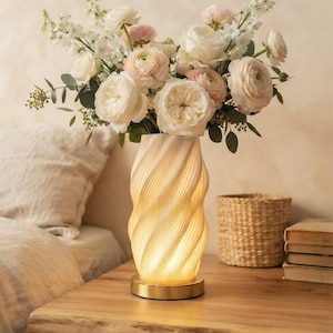 May include: A cylindrical table lamp with a white, spiraling, textured body that emits a warm, soft glow. The lamp base is a circular, metallic gold disc. A large bouquet of white and pale pink flowers is arranged inside the top of the lamp.