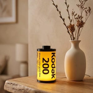 Puede incluir: Una lámpara cilíndrica diseñada para parecerse a un rollo de película fotográfica Kodak 200 de 35mm. La lámpara presenta un cuerpo amarillo con texto negro y tapas negras en la parte superior e inferior, emitiendo una luz cálida desde el interior.