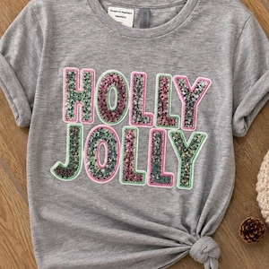 Può includere: Una t-shirt grigio melange a maniche corte con la scritta HOLLY JOLLY in grandi lettere in stile blocco. Le lettere sono riempite di paillettes verdi e rosa e bordate di verde chiaro. La maglietta presenta un orlo annodato in basso a destra.
