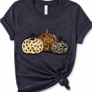 May include: A dark heather grey short-sleeve t-shirt featuring a graphic of three pumpkins. The left pumpkin is cream with a black leopard print, the centre is gold glitter, and the right is grey with a black leopard print. The shirt has a crew neckline.