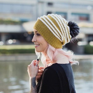 Op de afbeelding: Een vrouw met roze haar draagt een gebreide muts met gele en witte strepen en een zwarte pompon.