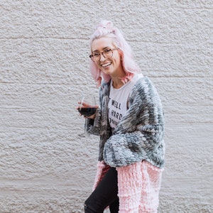 Może przedstawiać: Kobieta z różowymi włosami ma na sobie szary i różowy kardigan i trzyma kieliszek czerwonego wina. Uśmiecha się i patrzy w obiektyw. Tekst na jej koszulce brzmi "Postaram się dla ciebie".