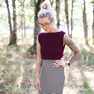Op de afbeelding: Een vrouw draagt een bordeauxrood gebreid crop top met een zwart-wit gestreepte rok. Ze heeft roze haar en draagt een bril.