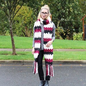 May include: A woman wearing a black and white striped scarf with burgundy accents and tassels. The scarf is crocheted and has a thick texture.